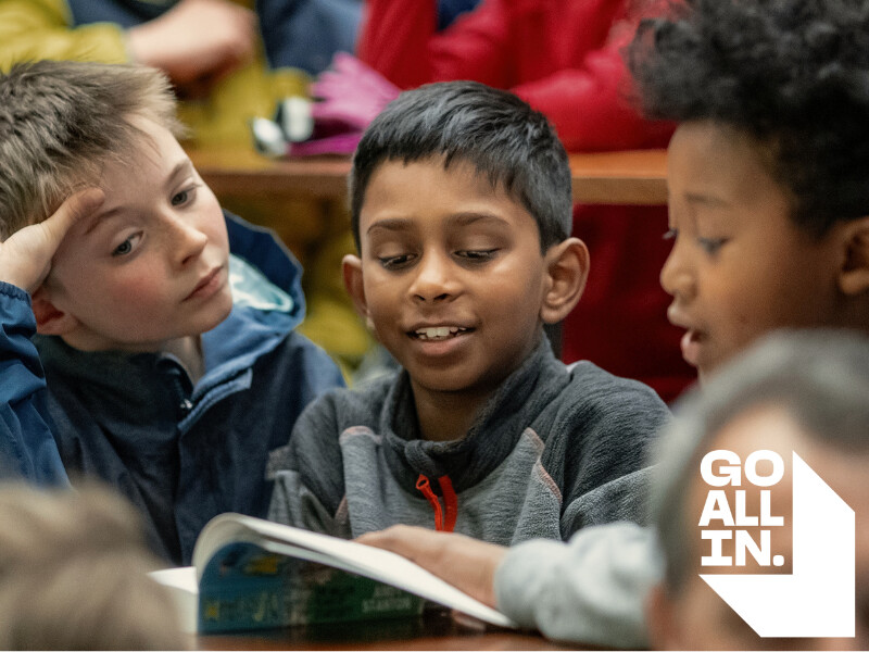 Three young boys reading together with a white logo in the corner that reads 'Go All In'